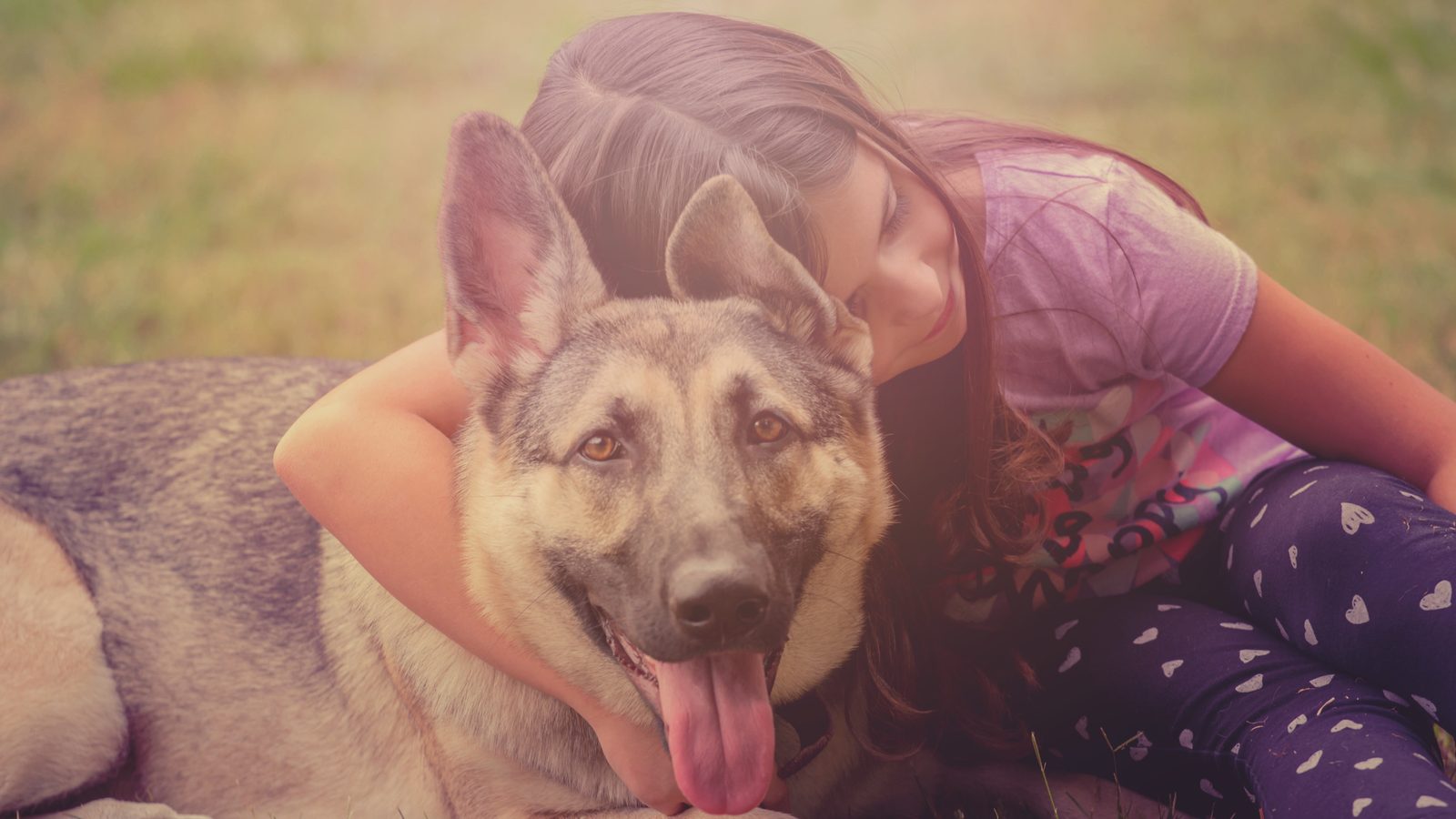 Ein Kind liegt neben einem großen Hund im Gras und umarmt ihn